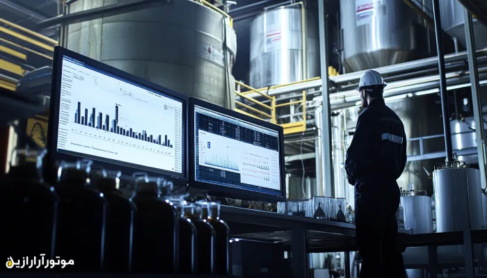 کارشناس فنی با کلاه ایمنی در حال بررسی دادههای تحلیلی روی دو مانیتور بزرگ در کارخانه تولید روانکار، با پسزمینهای از مخازن صنعتی و محیط نیمهتاریک پیشرفته.