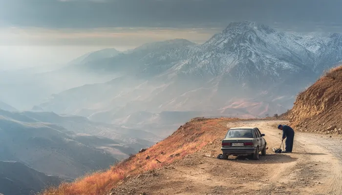خودرو قدیمی متوقف‌شده در جاده خاکی کوهستانی با منظره برفی و مکانیکی که در حال بررسی چرخ یا موتور است