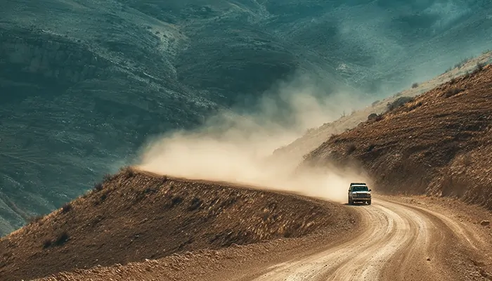 خودروی آفرود در حال حرکت روی جاده خاکی کوهستانی با گرد و غبار در پس‌زمینه و چشم‌انداز مرتفع