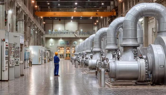 مهندس با کلاه ایمنی زرد در سالن بزرگ نیروگاه یا ایستگاه پمپاژ صنعتی، در میان ردیف لوله‌ها و توربین‌های نقره‌ای